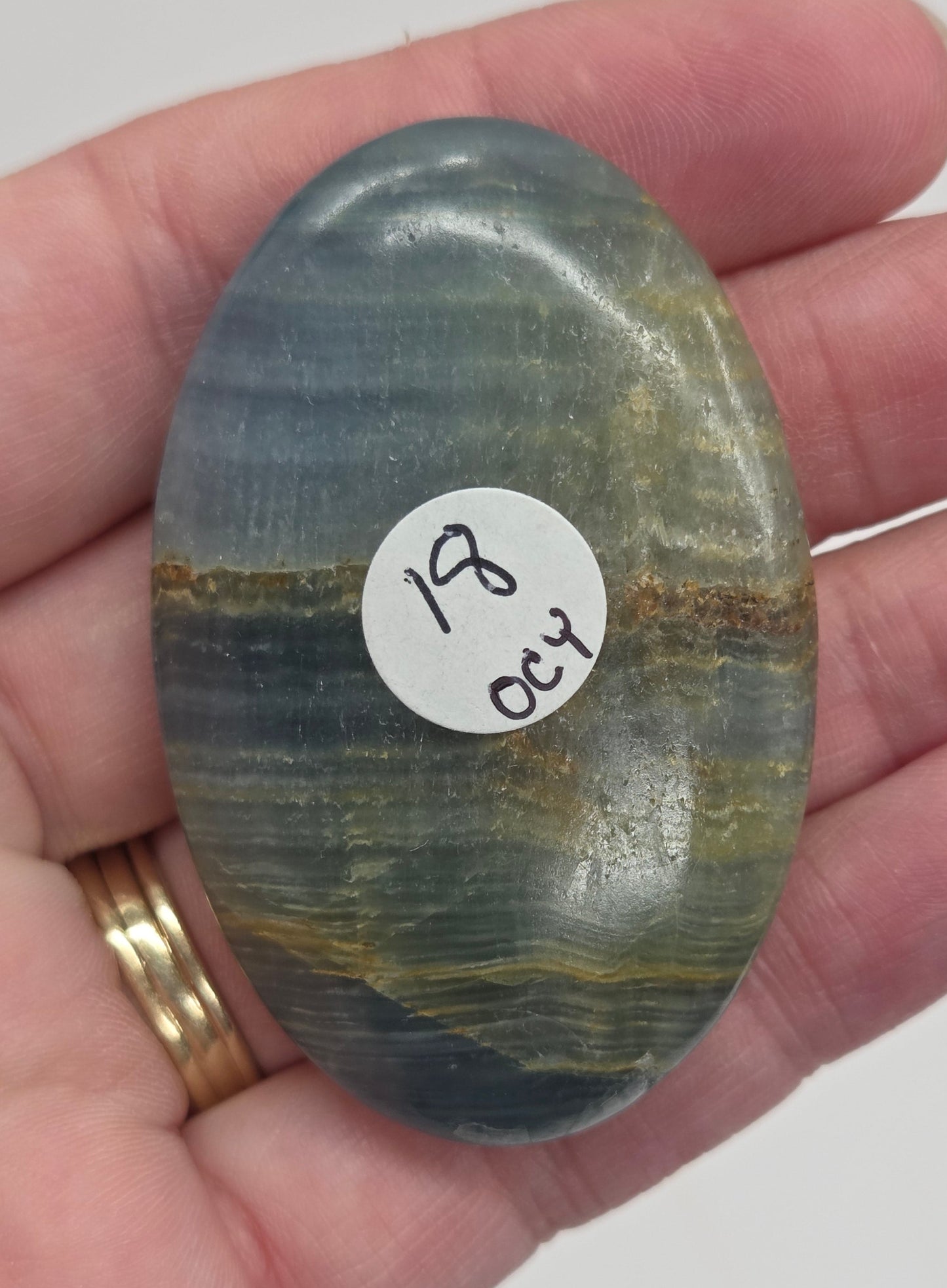 Hand holding a smooth, oval-shaped Blue Onyx palm stone with banded patterns of blue, white, and brown.