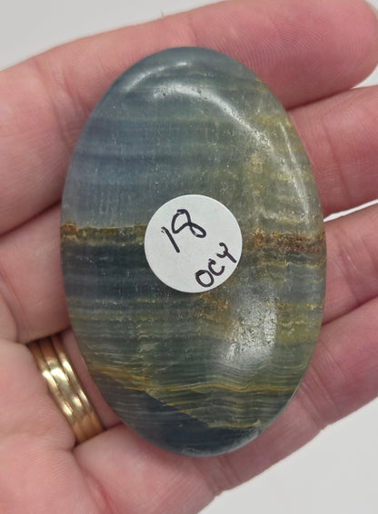 Hand holding a smooth, oval-shaped Blue Onyx palm stone with banded patterns of blue, white, and brown.