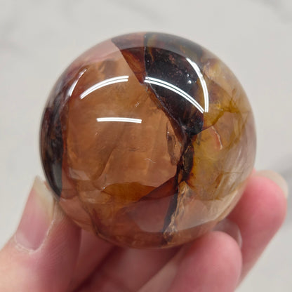 Hand holding a polished sphere with a mix of golden healer and fire quartz  against a white background