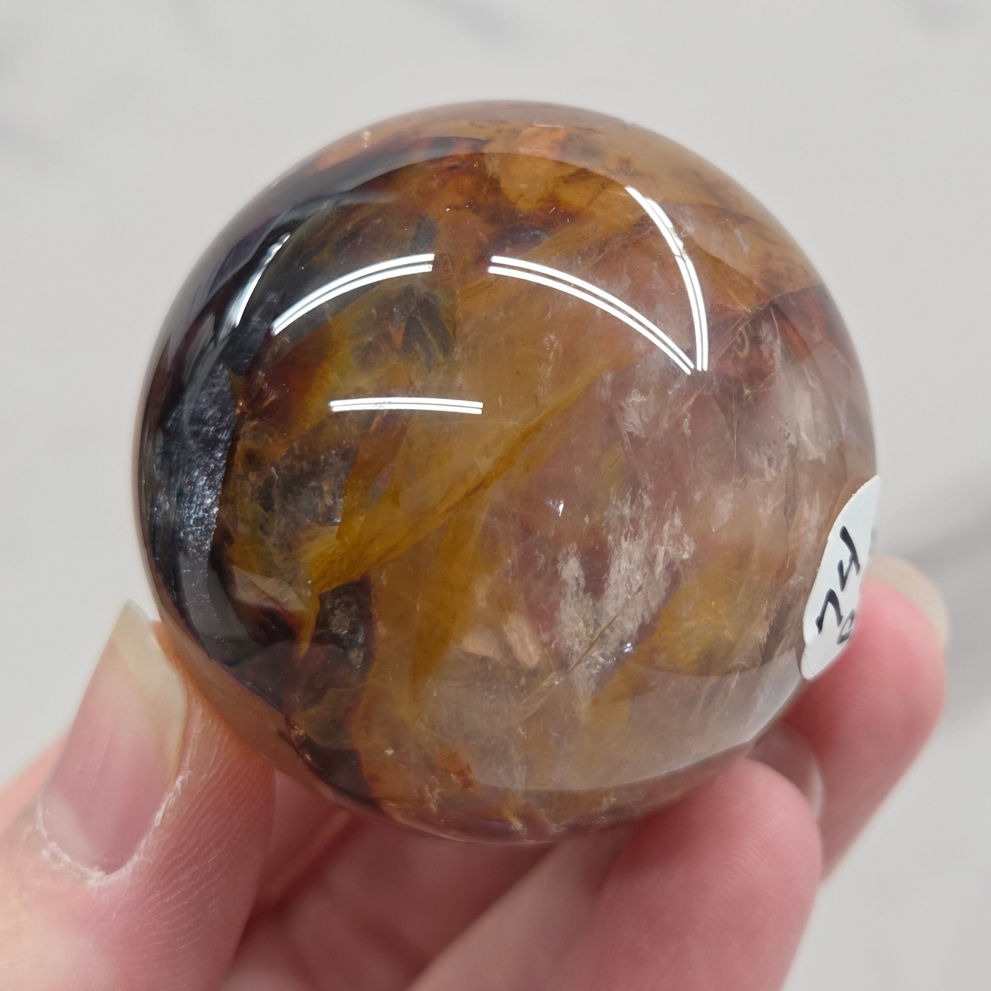 Hand holding a polished sphere with a mix of golden healer and fire quartz  against a white background