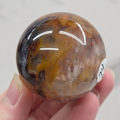 Hand holding a polished sphere with a mix of golden healer and fire quartz  against a white background