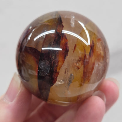 Hand holding a polished sphere with a mix of golden healer and fire quartz  against a white background