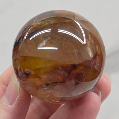 Hand holding a polished sphere with a mix of golden healer and fire quartz  against a white background