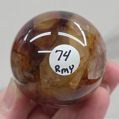 Hand holding a polished sphere with a mix of golden healer and fire quartz  against a white background