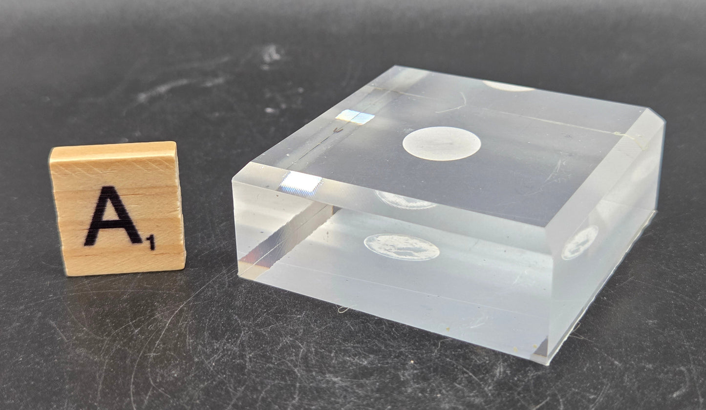 Clear square block with circular cutout next to a wooden letter 'A' on a dark surface