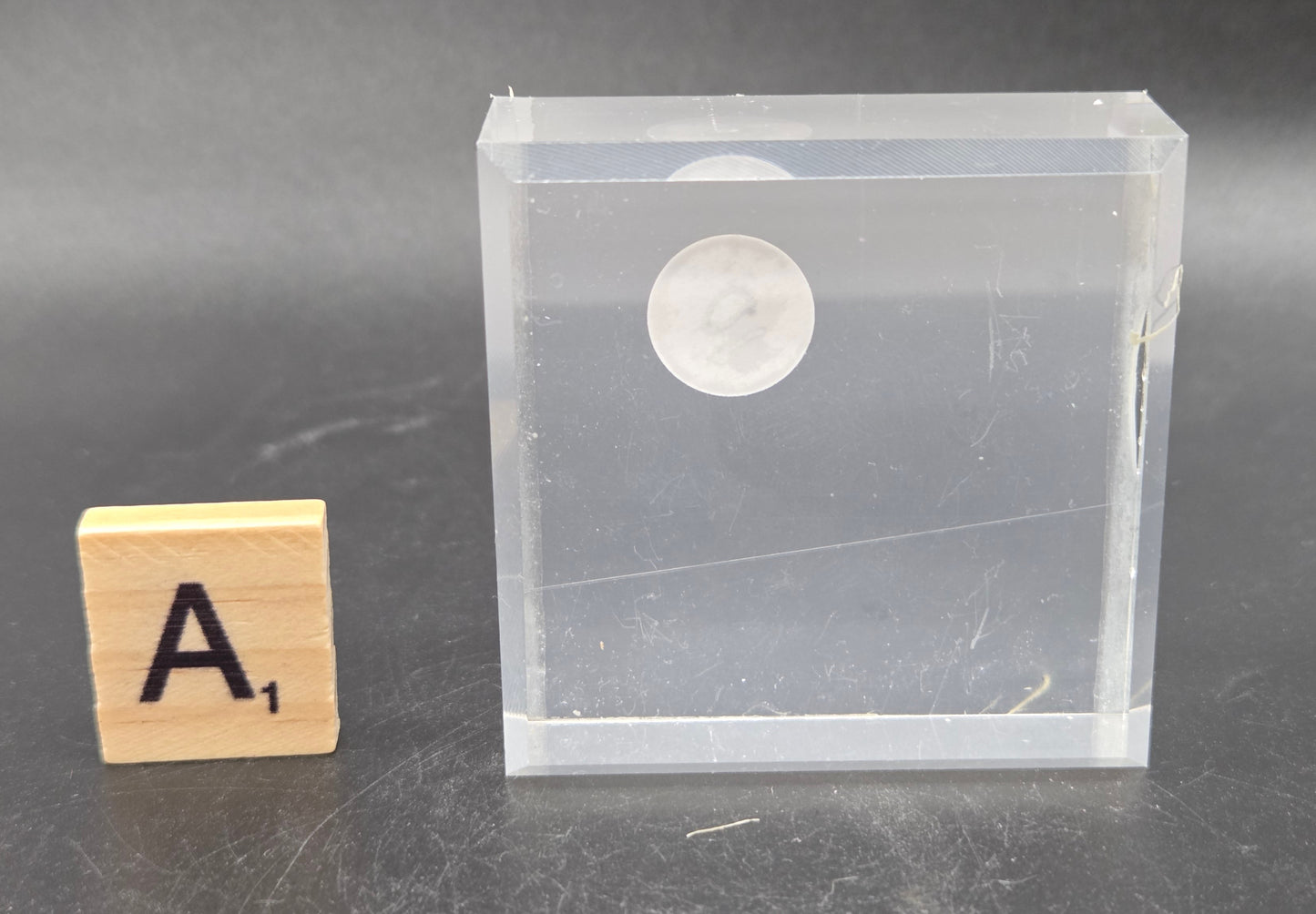 Clear plastic case with a circular object on a gray surface, next to a wooden block with the letter 'A'.