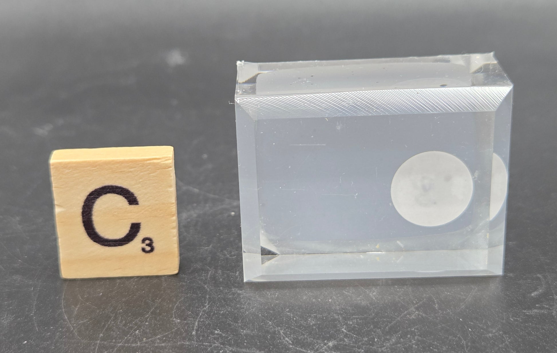 Clear rectangular block with circular component on a gray surface next to a wooden block with 'C' and '3' printed on it.