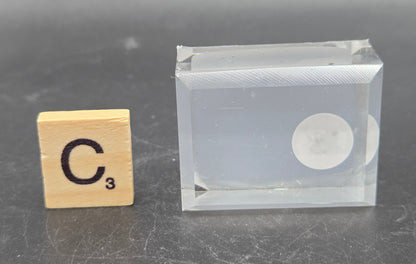 Clear rectangular block with circular component on a gray surface next to a wooden block with 'C' and '3' printed on it.