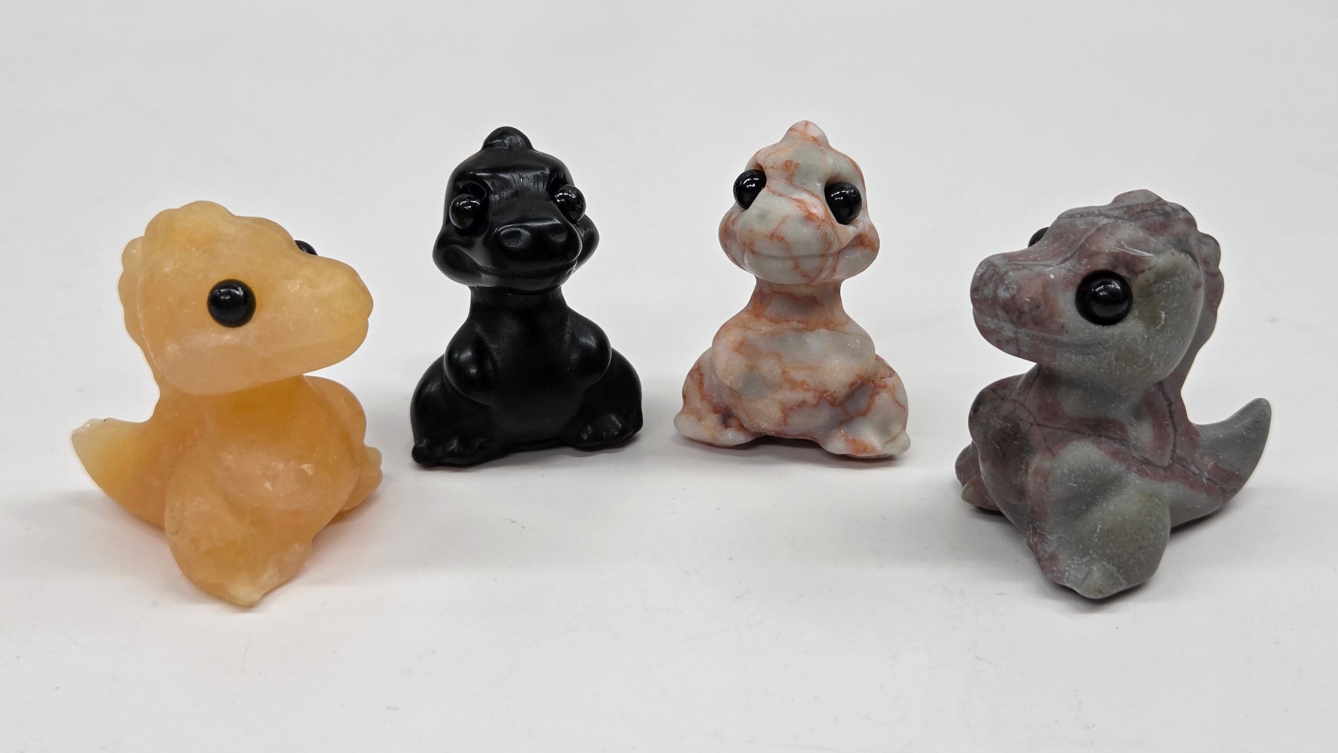 Four small baby t-rex dinosaur carvings in different colors on a white surface
