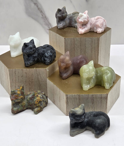 Cat carvings lying down in a variety of materials and colors on a wooden stand against a neutral background