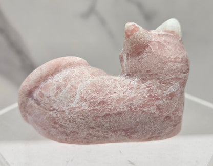 Pink Opal crystal cat carving lying down an acrylic stand against a neutral background