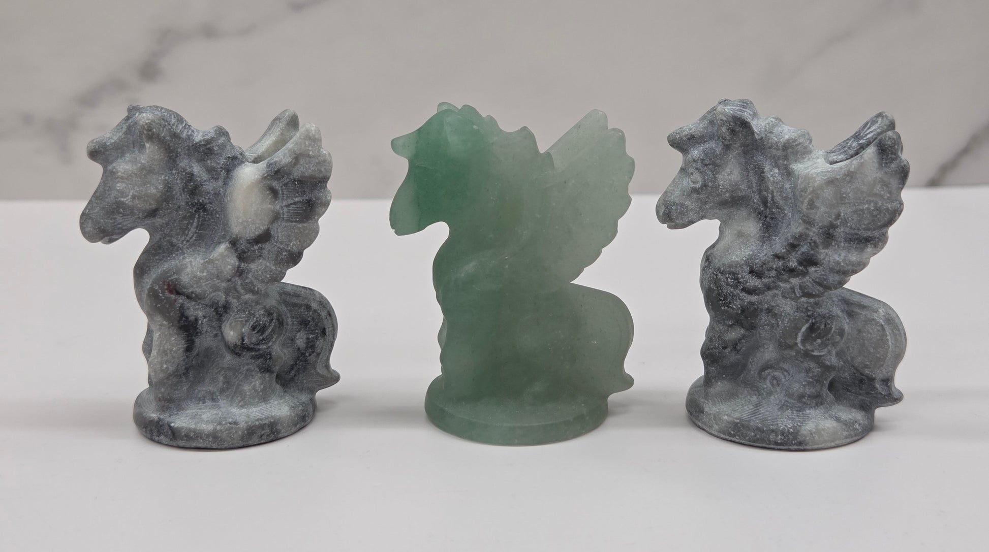 Three stone-like figurines of winged horses on a white surface with a marble-patterned wall in the background.