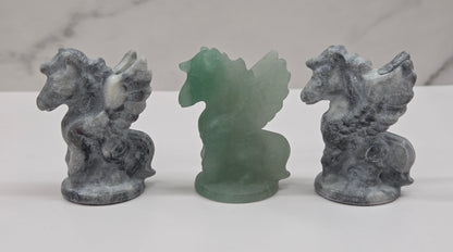 Three stone-like figurines of winged horses on a white surface with a marble-patterned wall in the background.