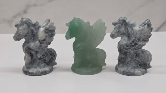 Three stone-like figurines of winged horses on a white surface with a marble-patterned wall in the background.