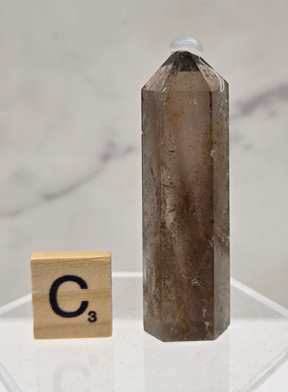 Smoky Quartz crystal on a clear stand with a letter 'C' block against a light background