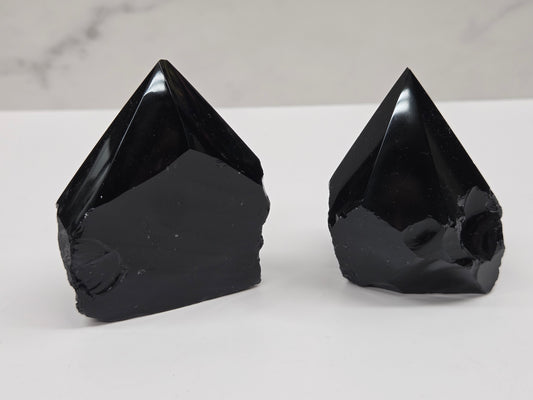 Two black Obsidian half polished points on a white surface with a gray marble background