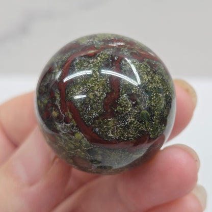 Hand holding a dragons blood sphere  with green and red patterns against a plain background