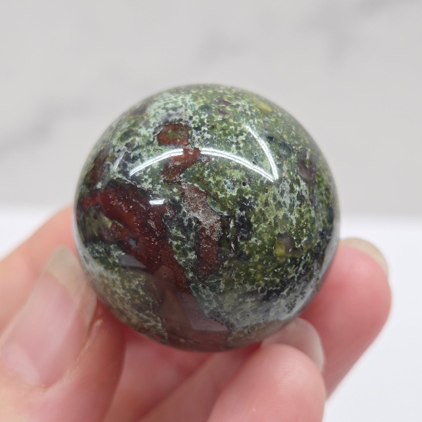 Hand holding a dragons blood sphere  with green and red patterns against a plain background