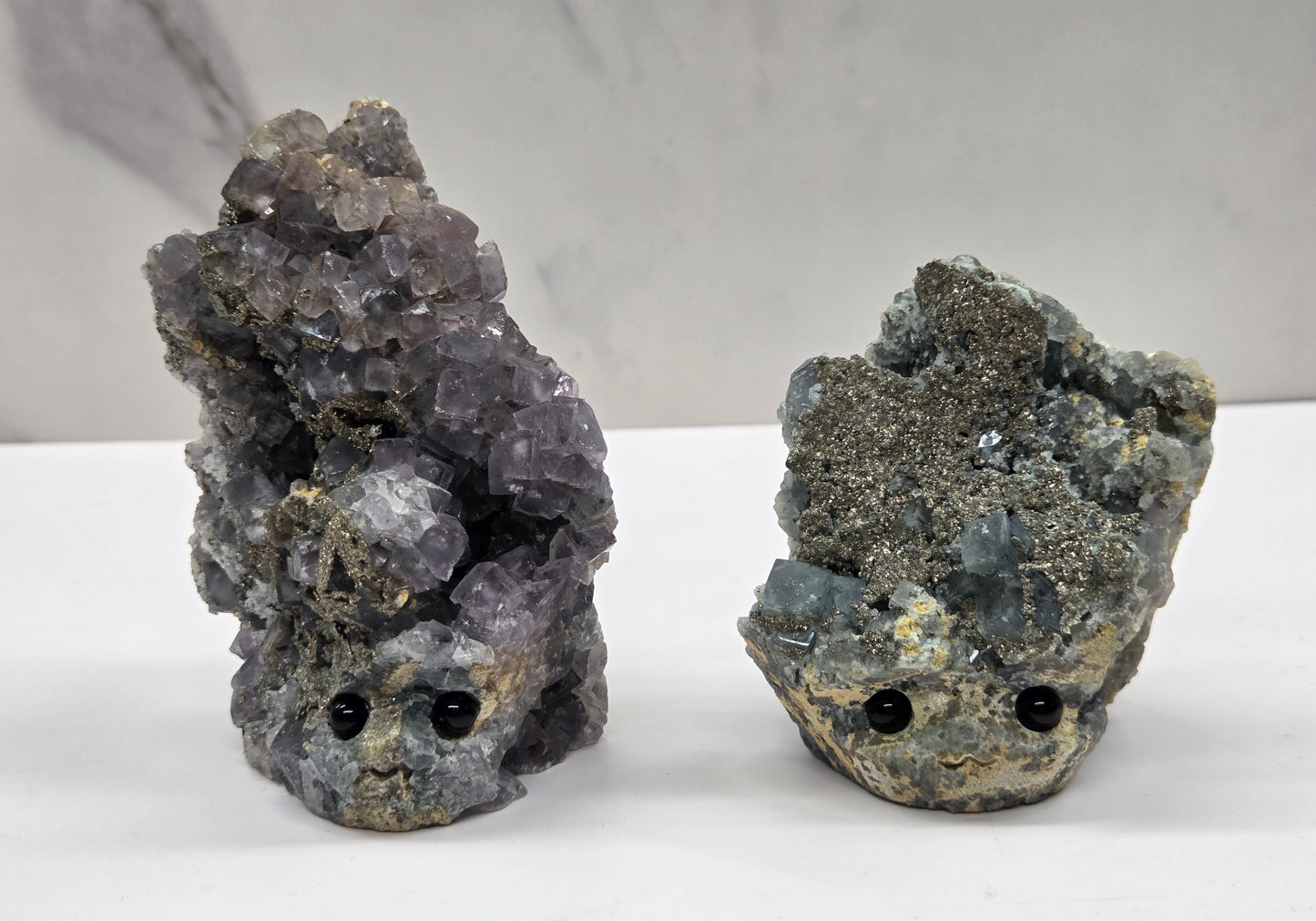 Two fluorite figurines with black eyes on a white surface and marble background