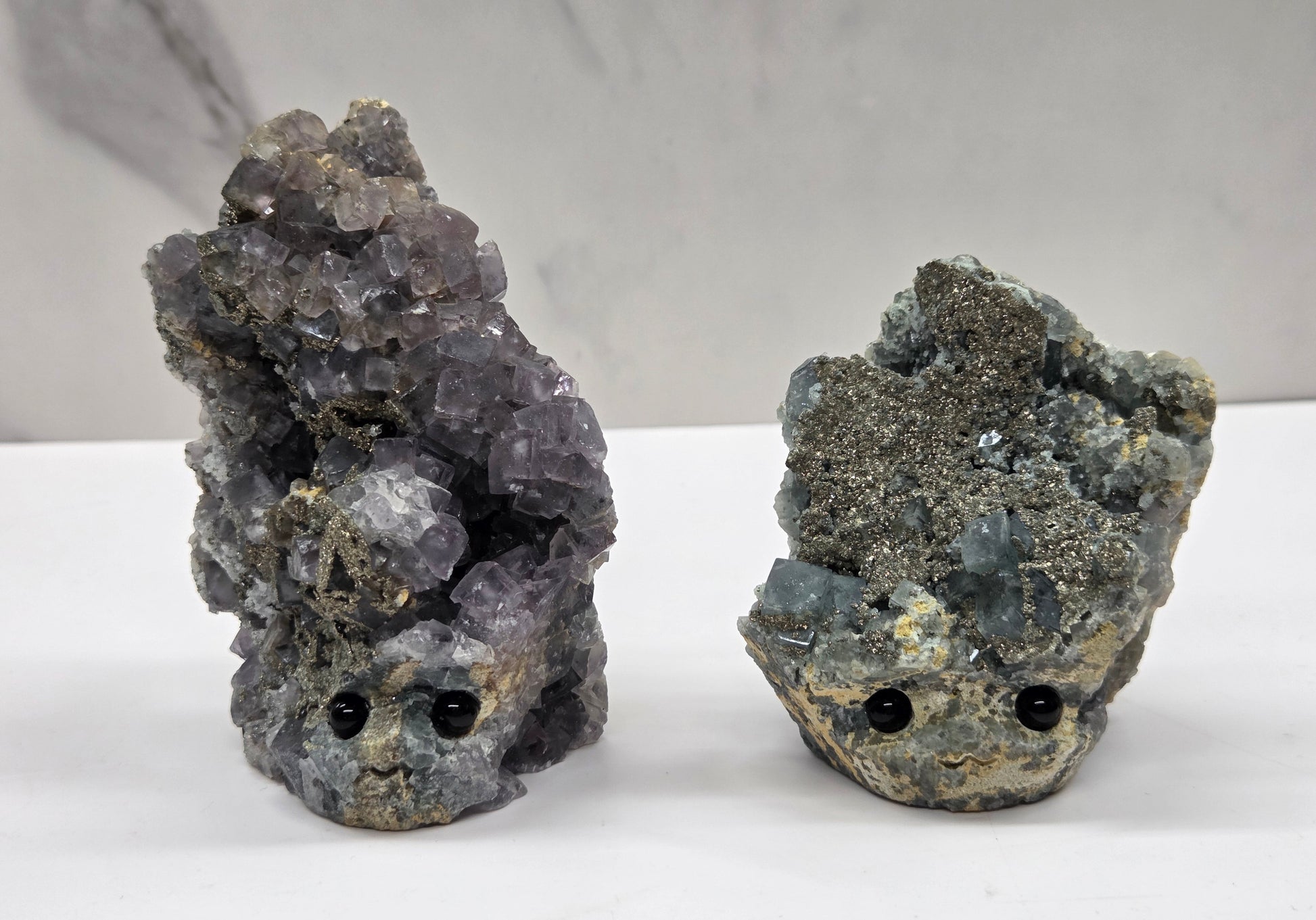 Two fluorite figurines with black eyes on a white surface and marble background