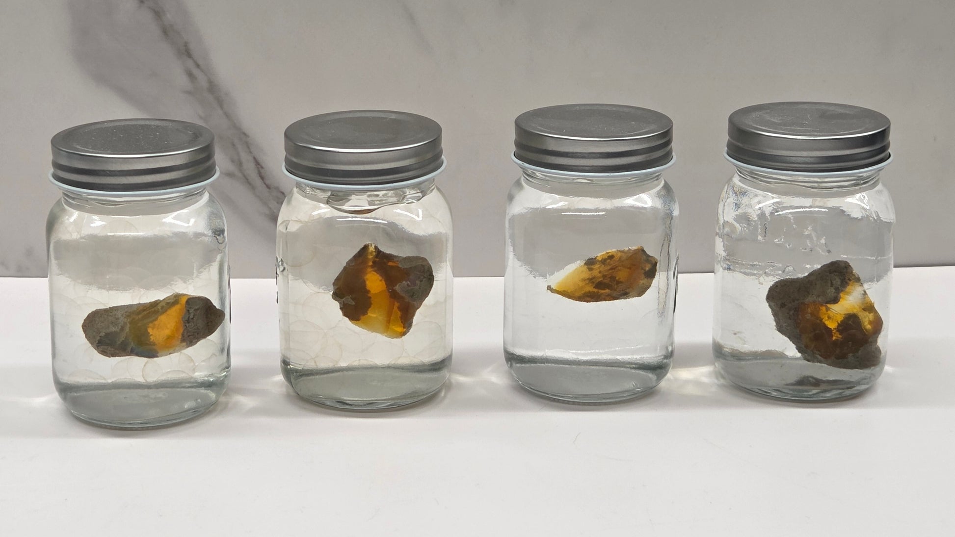 Four glass jars with metal lids containing opals on a marble surface.