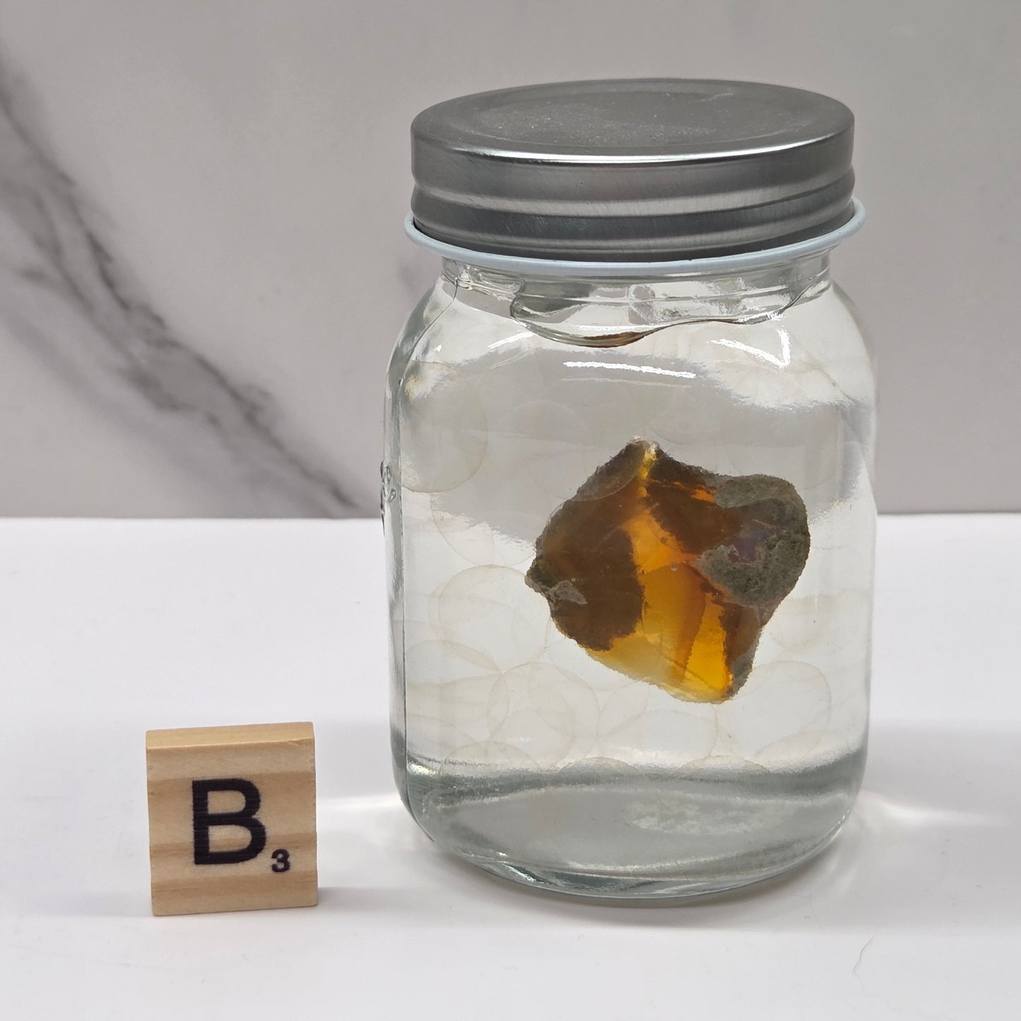 Clear jar with a metallic lid containing a brown rock, on a white surface with a marble-patterned wall in the background.