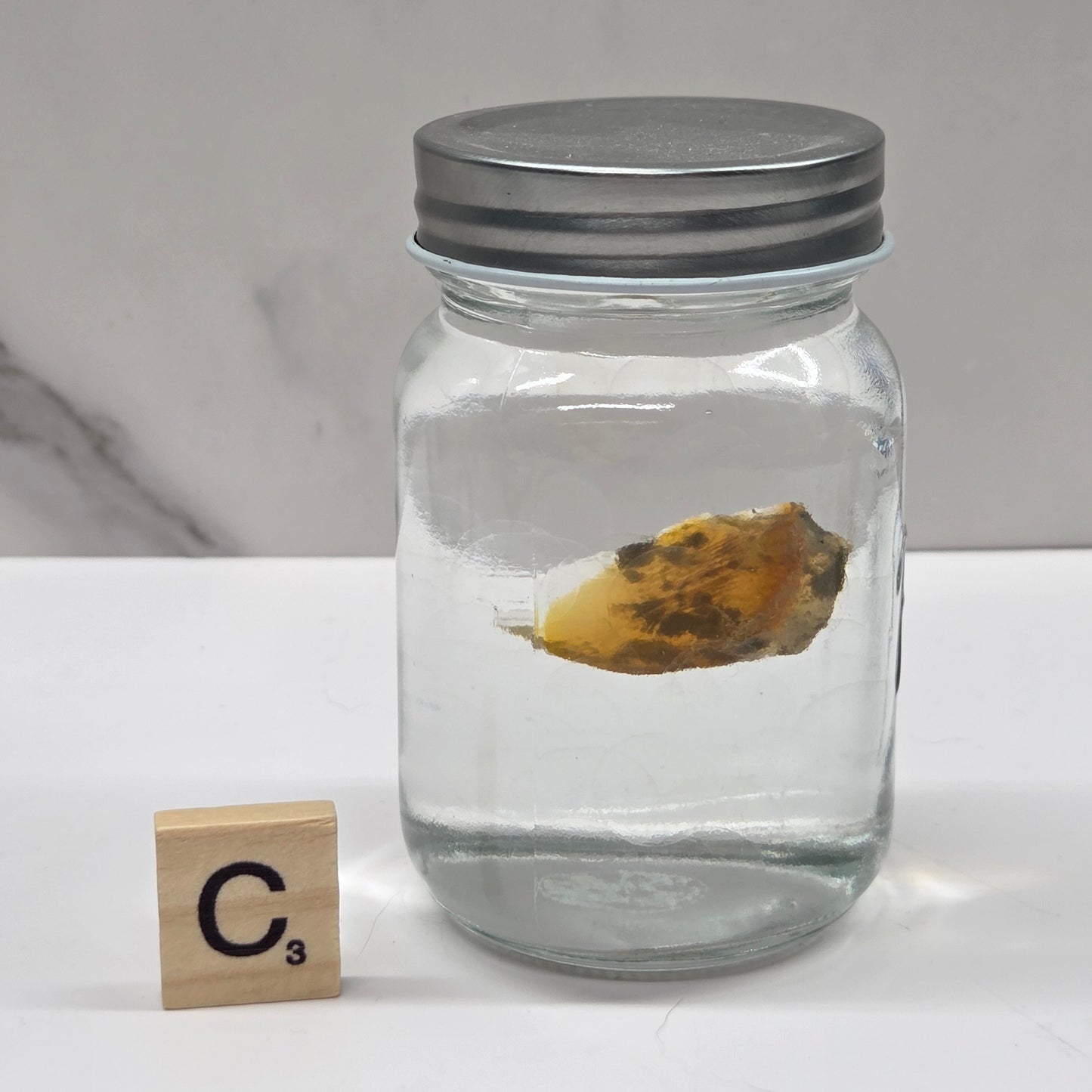 Clear glass jar with a metal lid containing a yellowish object on a white surface.