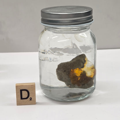 Clear jar with a metal lid containing a rock, on a white surface with a wooden block labeled 'D2'.