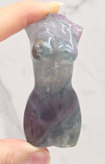 Female torso carving made of fluorite  being held between fingers on a white background