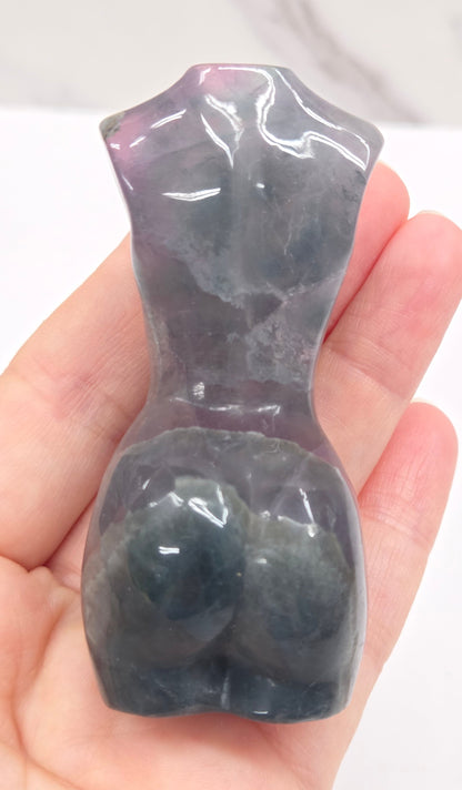 Female torso carving made of fluorite being held in a hand on a white background
