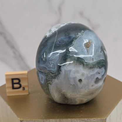 Moss Agate skull carving on a wood stand against a white background