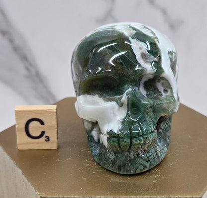 Moss Agate skull carving on a wood stand against a white background