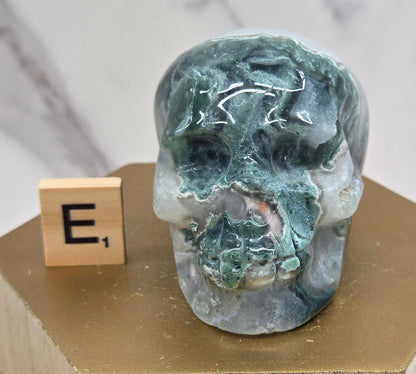 Moss Agate skull carving on a wood stand against a white background
