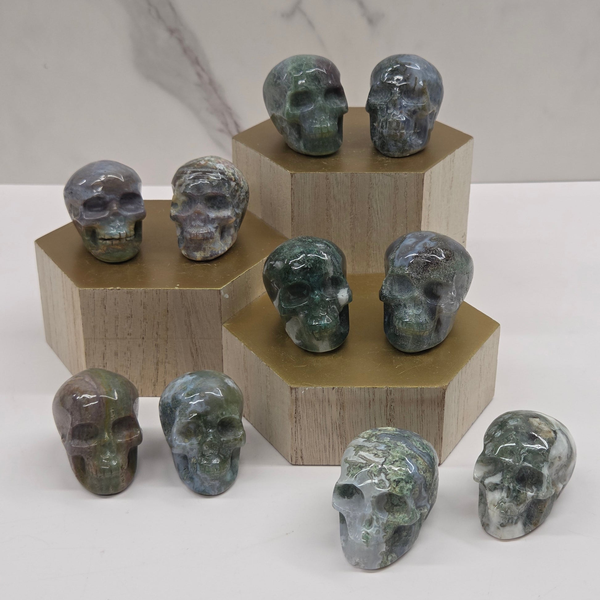 A collection of medium-sized skull-shaped rocks displayed in a clear, three-tiered stand, with varying shades of green and white, made of Moss Agate