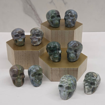 A collection of medium-sized skull-shaped rocks displayed in a clear, three-tiered stand, with varying shades of green and white, made of Moss Agate