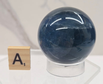 A polished dark blue fluorite spherical stone displayed on a clear acrylic stand next to a wooden Scrabble tile showing the letter A.