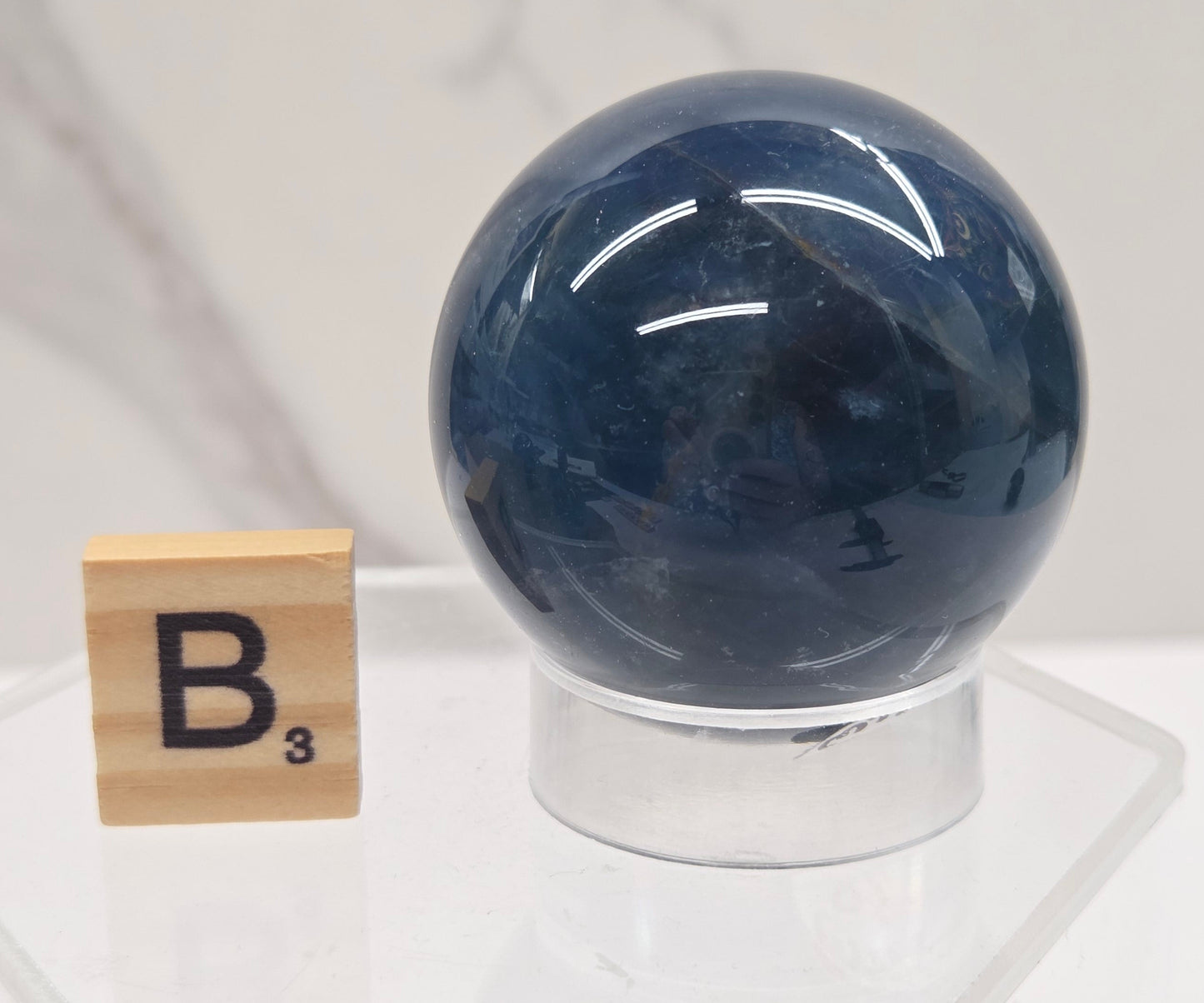 A polished dark blue fluorite crystal sphere mounted on a clear acrylic stand with a small wooden periodic table tile displaying 'B³' underneath, set against a white marble background.