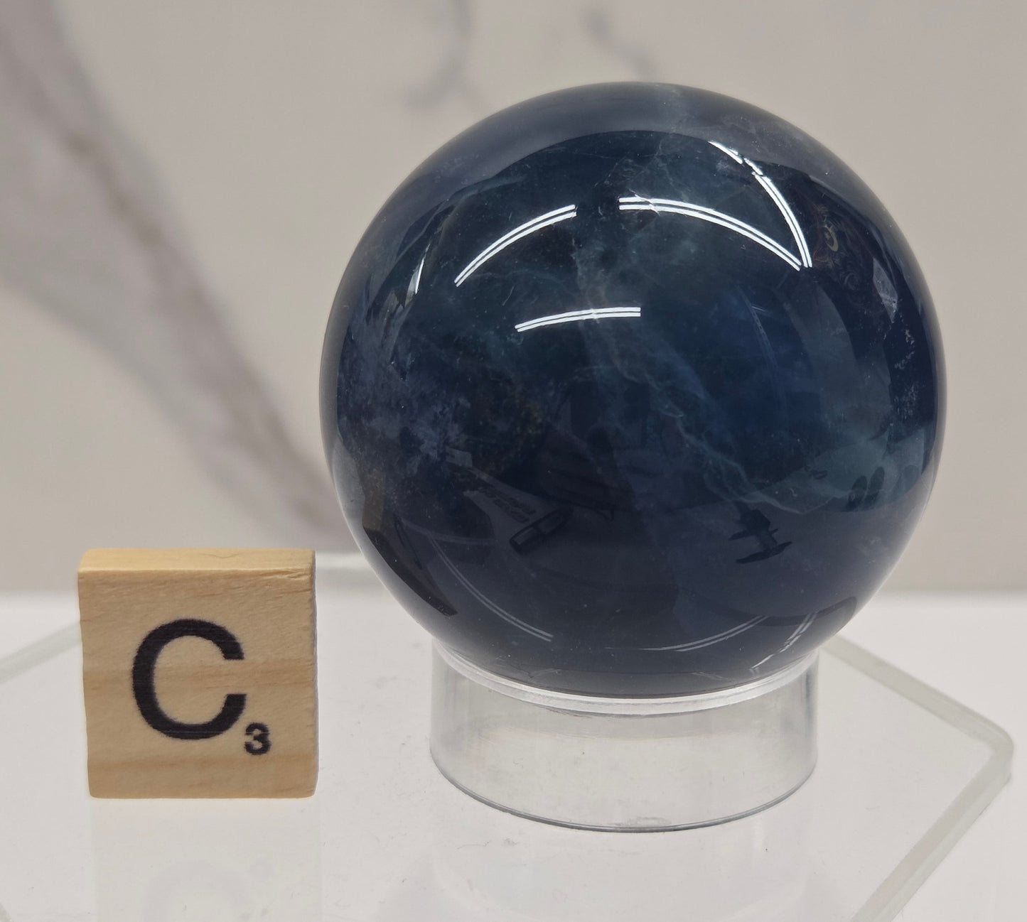 A polished dark blue spherical crystal mounted on a clear acrylic stand, with a small wooden tile marked C³ placed below it against a light marble-like background.