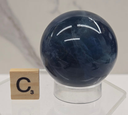 A polished dark blue spherical crystal mounted on a clear acrylic stand, with a small wooden tile marked C³ placed below it against a light marble-like background.