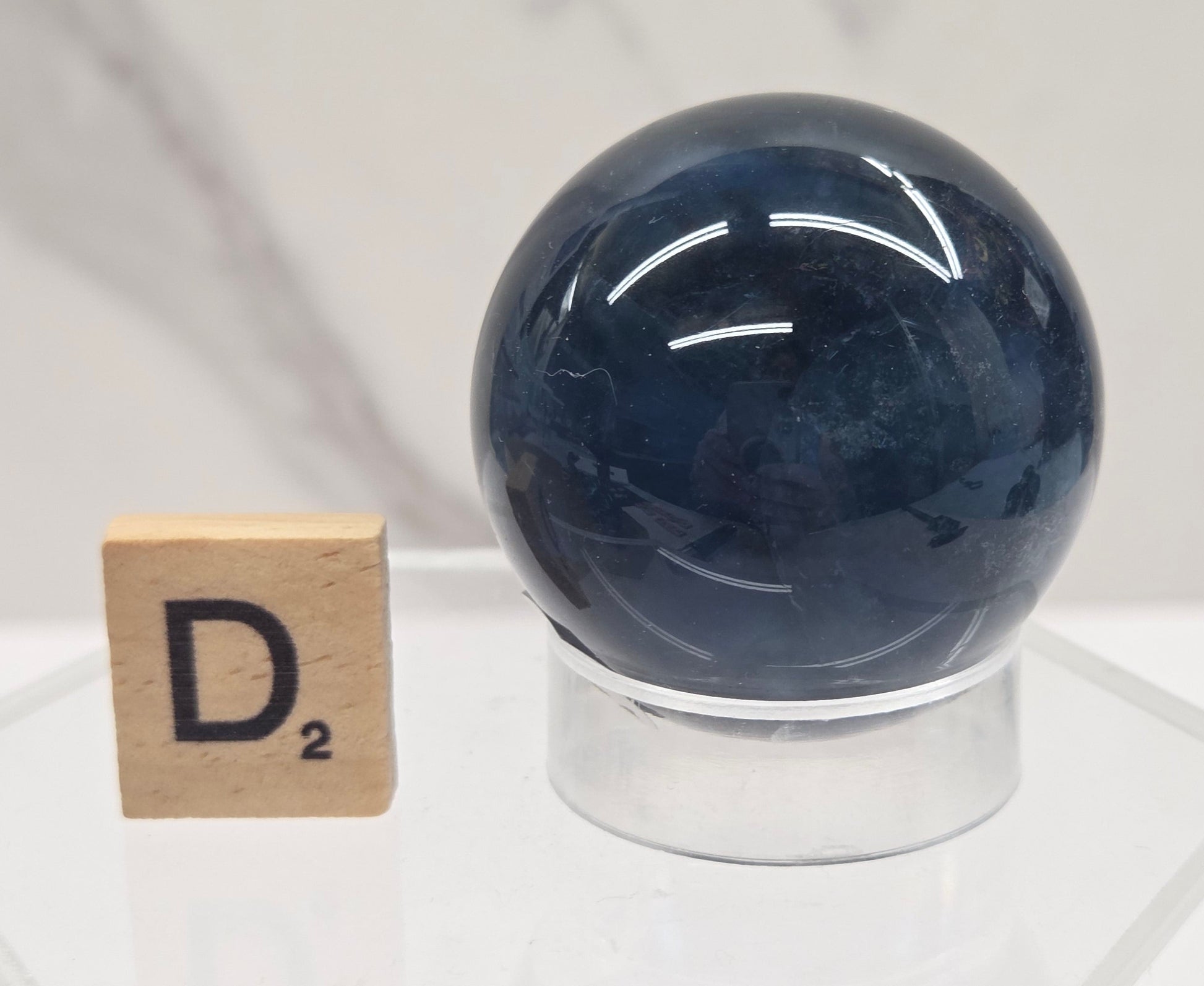 A polished dark blue gemstone sphere mounted on a clear acrylic stand, with a small wooden letter tile showing the letter D nearby, set against a light marble-patterned background.