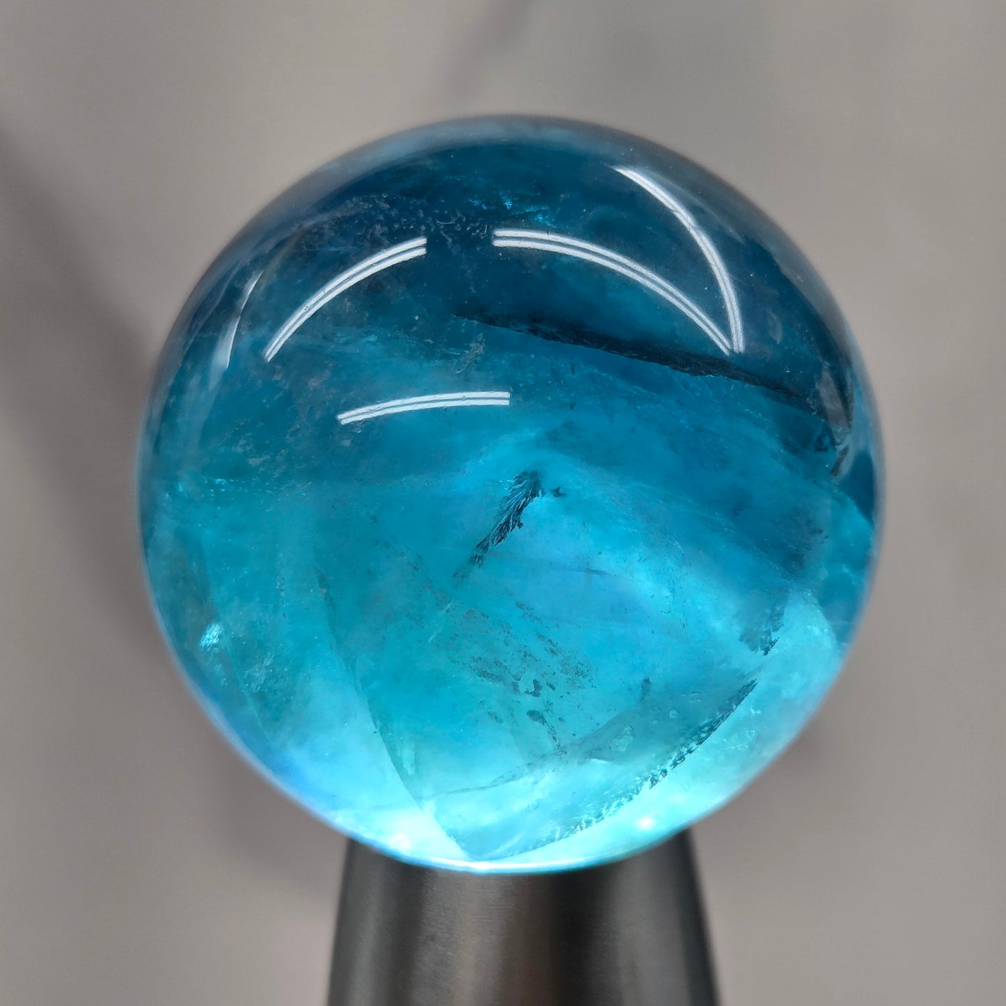 A close-up of a polished, spherical blue gemstone held by a dark metal stand against a light gray marble background.