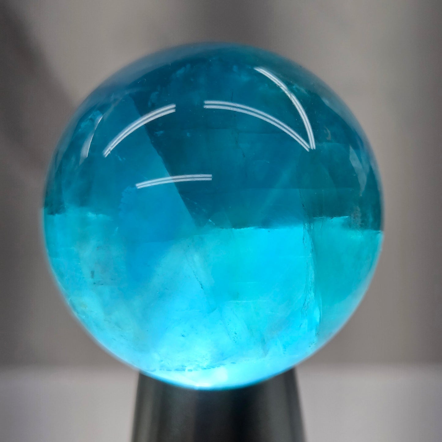 A close-up of a polished blue gemstone sphere held by a black tool, against a blurred gray background.