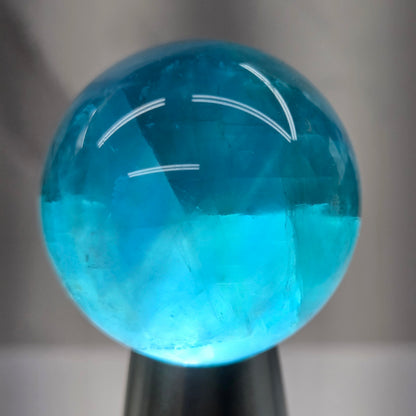 A close-up of a polished blue gemstone sphere held by a black tool, against a blurred gray background.