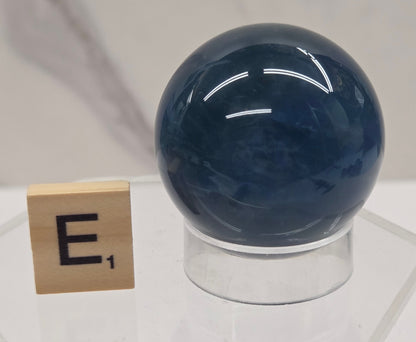 A polished dark blue spherical object mounted on a clear acrylic stand, with a small gold block displaying the Hebrew letter Shin below it, set against a light marble background.