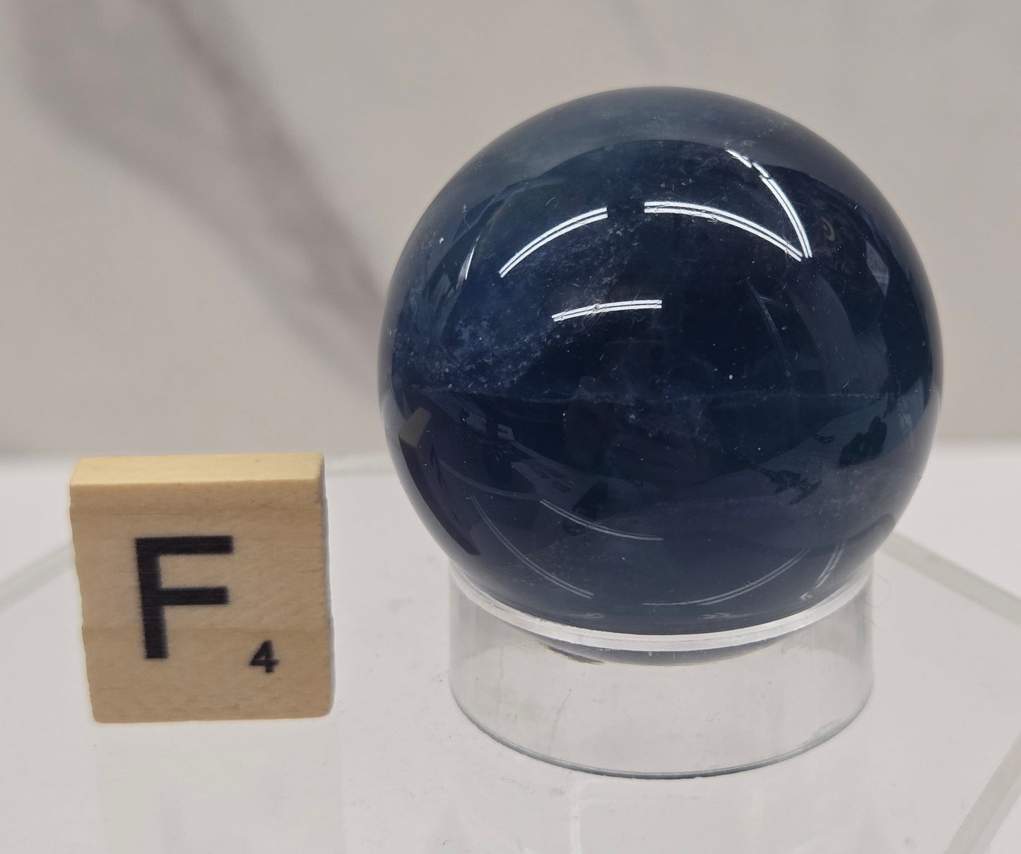 A dark blue spherical object mounted on a clear acrylic stand, with a small wooden tile labeled 'F' placed below it against a light marble-like background.
