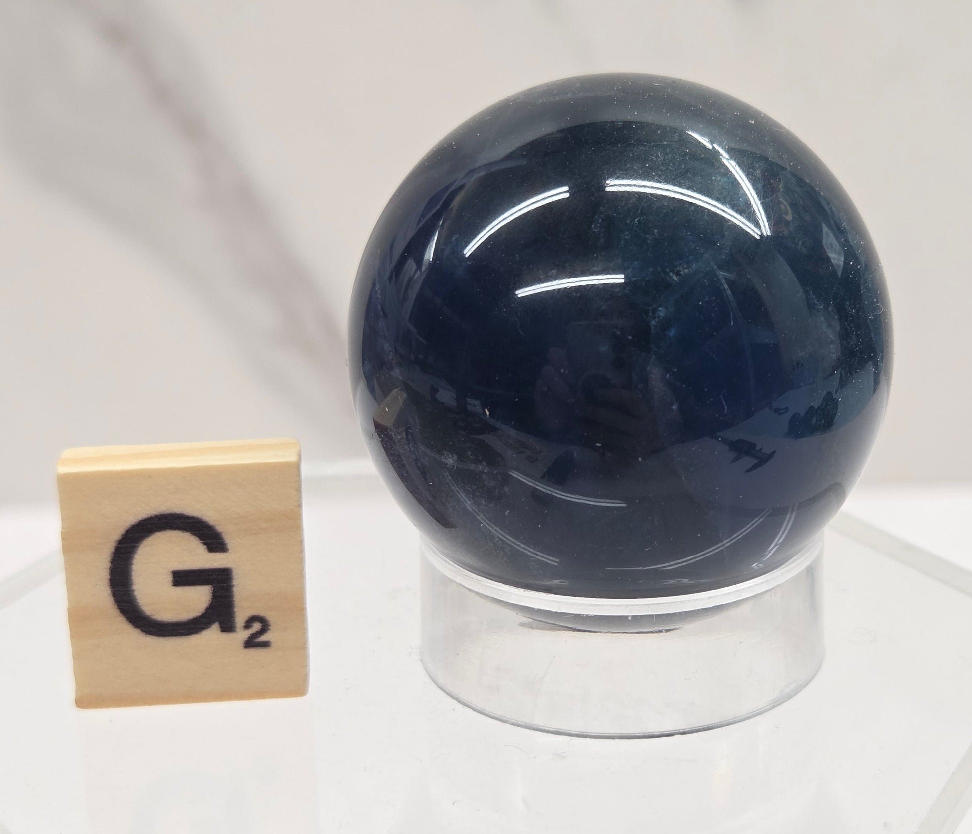 A dark blue spherical object mounted on a clear acrylic stand, with a small wooden tile labeled 'G' placed below it against a light marble-like background.