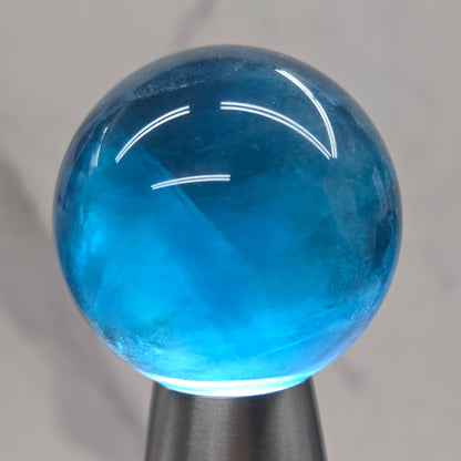 A close-up of a hand holding a metallic silver cylindrical object with a translucent blue spherical end against a light marble background.