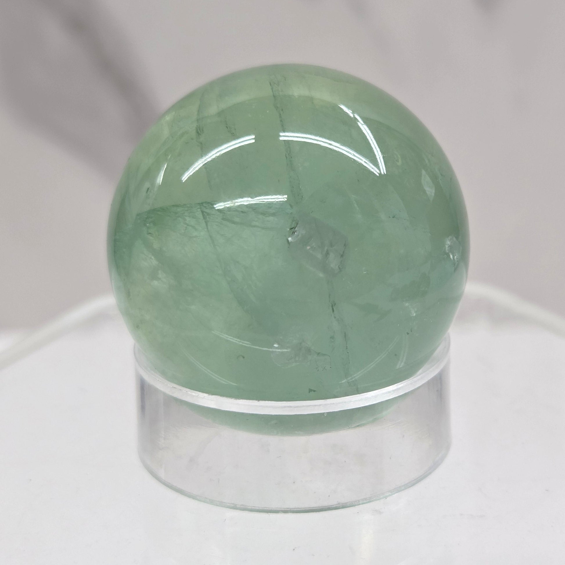 A close-up of a round, polished light green fluorite sphere mounted on a clear acrylic stand against a white marble-like background.
