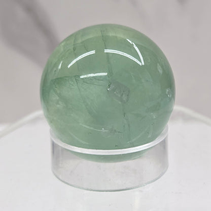 A close-up of a round, polished light green fluorite sphere mounted on a clear acrylic stand against a white marble-like background.