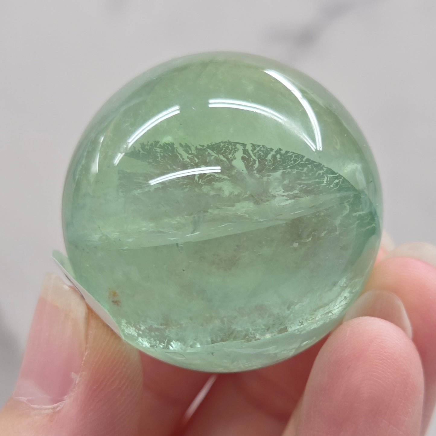 A close-up of a round, polished light green fluorite sphere mounted on a clear acrylic stand against a white marble-like background.